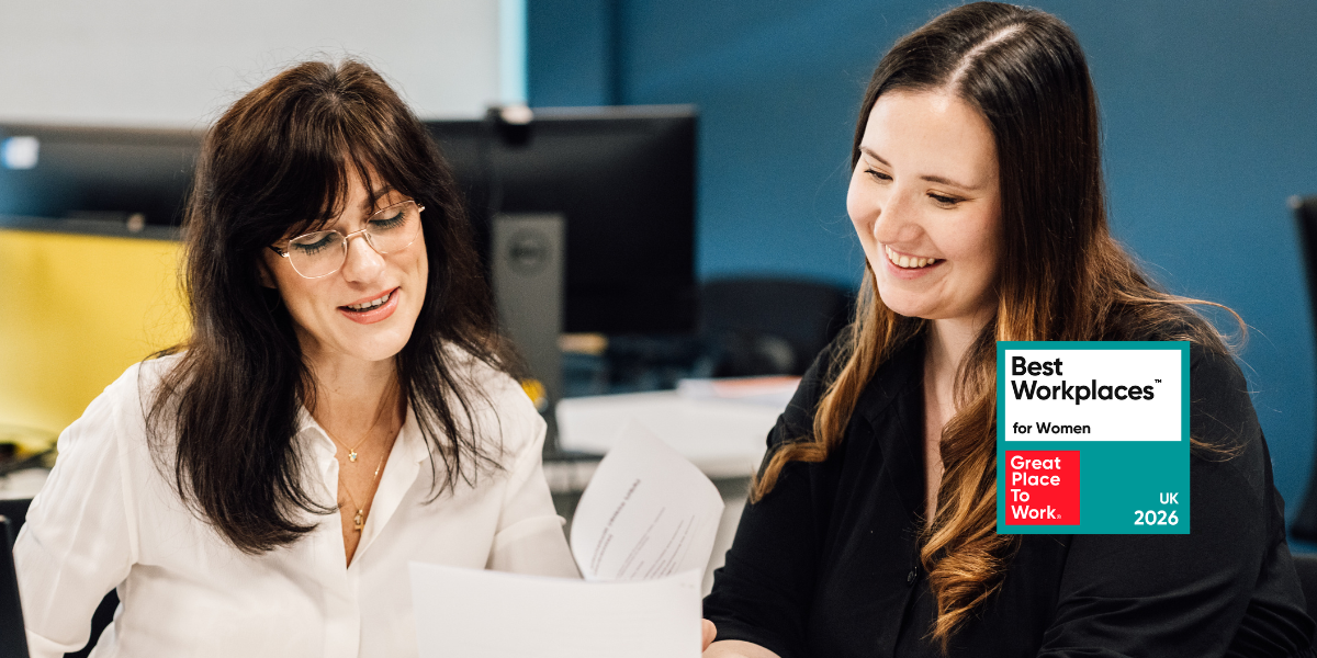 Two women sat at a desk reviewing a piece of paper. Great Place to Work logo for UK's Best Workplaces for Women placed to the right